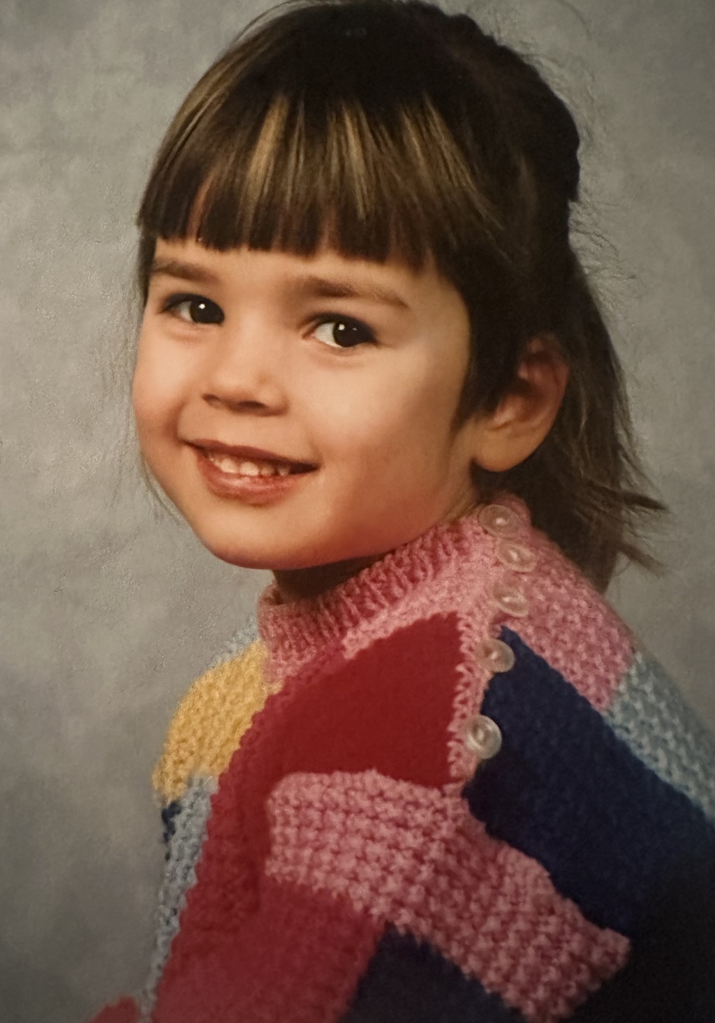 Koulukuva tai päiväkotikuva, jossa nuori tyttö istuu sivullepäin, kylki kohti kameraa. Päällä monivärinen neule, tyttö hymyilee ja katsoo kameraa valloittavasti.  